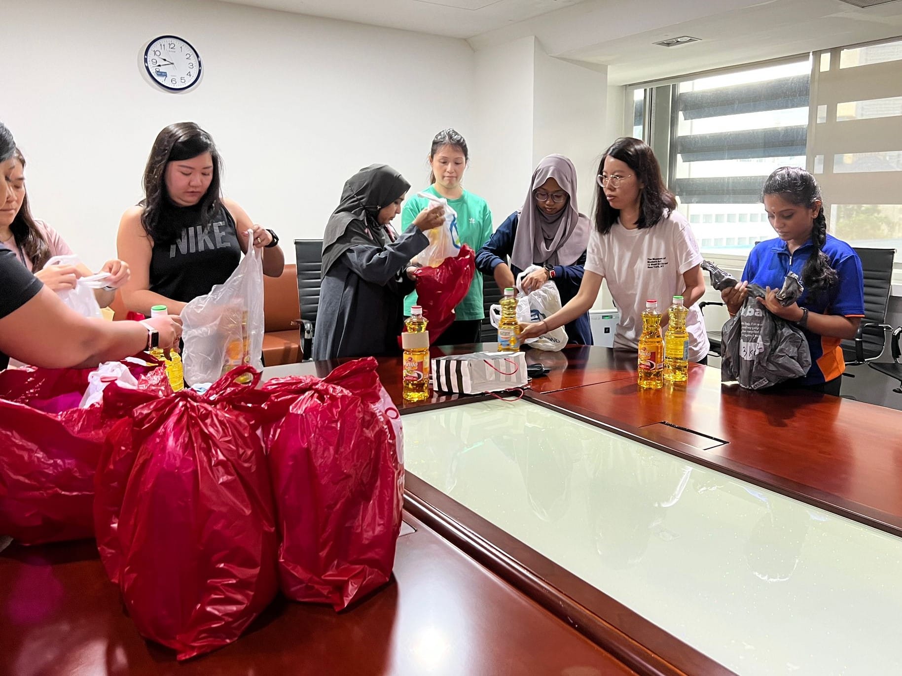 Six young women pack red bags with cooking oil bottles on a conference room table under a wall clock.